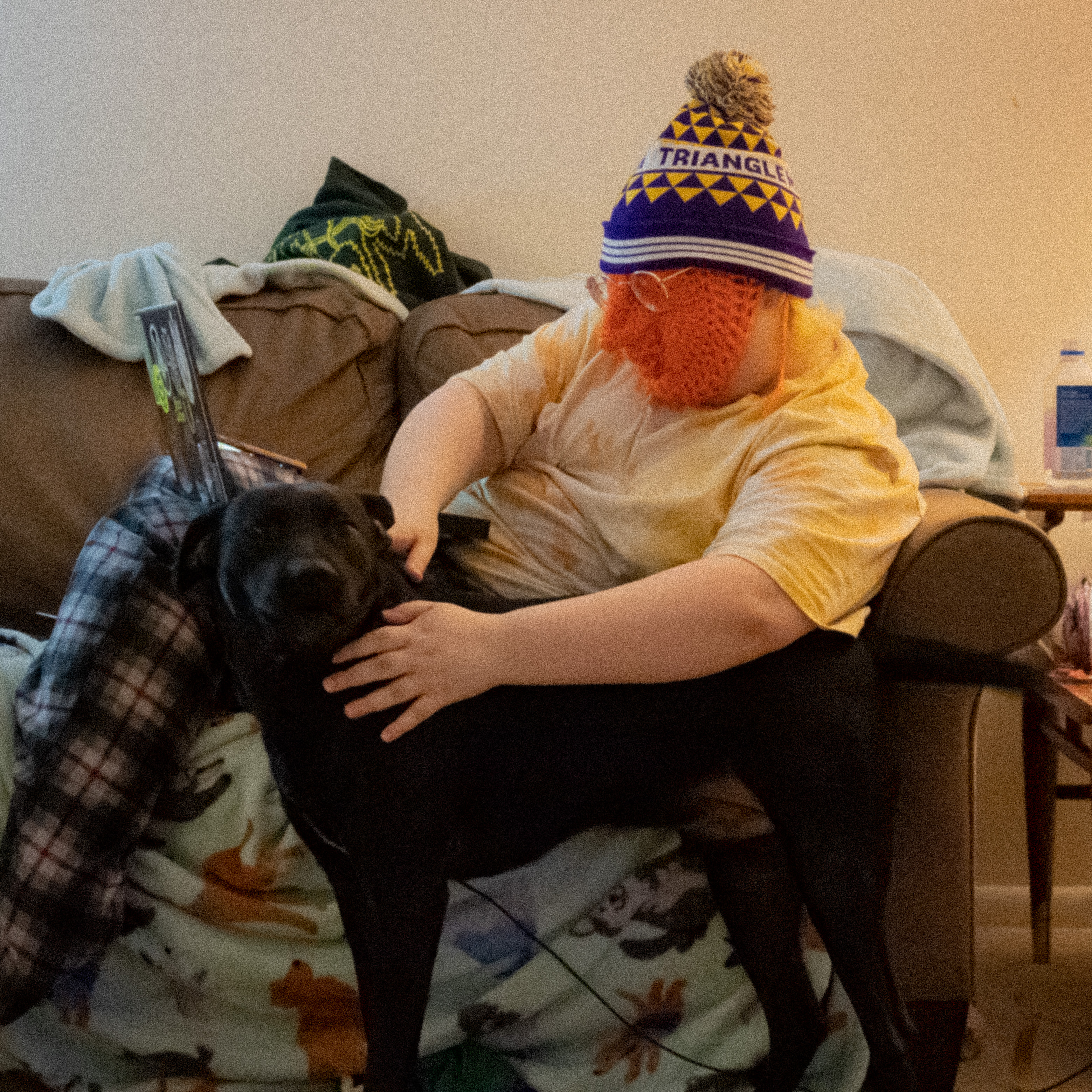 A caucasian person reclines on a couch, wearing an orange mask that conforms to their face and a festive winter hat. They are petting a dog, who looks smugly at the camera